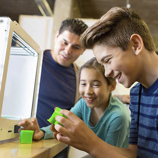 Preteen and elementary age siblings are using a 3D printer with their father in a makerspace studio. Family is printing 3D plastic objects with a cube style 3D printer. Mid adult man is watching his children learn about modern technology.